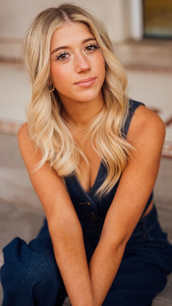 A young woman with long, wavy blonde hair and sun-kissed skin sits outdoors, wearing a sleeveless denim jumpsuit. She looks at the camera with a slight, soft smile and relaxed expression. by Justin Salem Meyer
