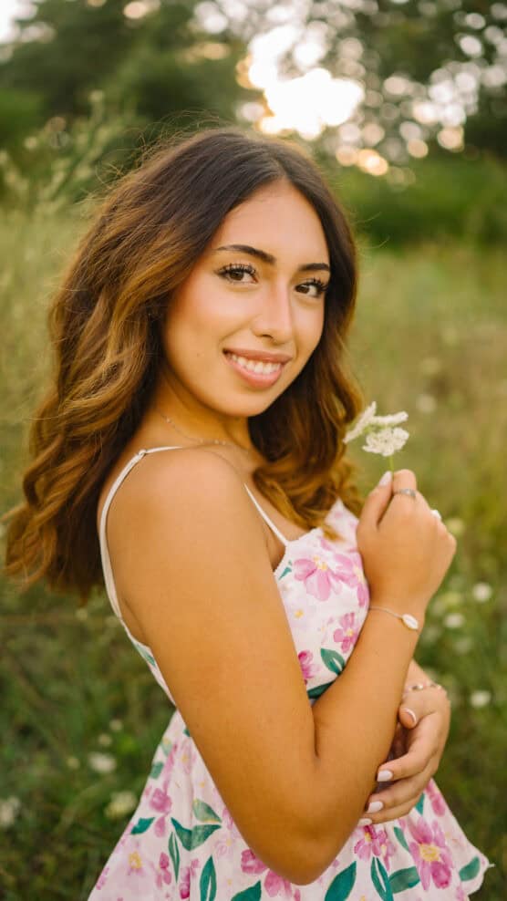 A young woman with wavy brown hair smiles while holding a small white flower. She wears a white floral dress and stands outdoors in a grassy, sunlit field with trees in the background. by Justin Salem Meyer