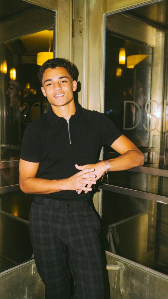 A young man in a black shirt and plaid pants stands in front of brass-framed glass doors, smiling with hands clasped. Warm indoor lights and reflections are visible in the glass behind him. by Justin Salem Meyer