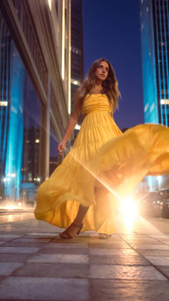 A woman in a flowing yellow dress walks confidently on a city sidewalk at night, illuminated by bright lights and surrounded by tall buildings with blue lighting. by Justin Salem Meyer