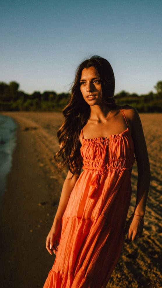 A woman with long wavy hair in an orange sundress stands on a sandy beach at sunset, looking to the side with greenery and trees in the background. by Justin Salem Meyer