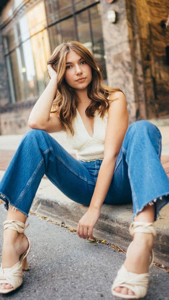 A young woman with long brown hair sits on a sidewalk, wearing a white sleeveless top, blue jeans, and cream-colored heeled sandals, looking at the camera with a relaxed expression. by Justin Salem Meyer