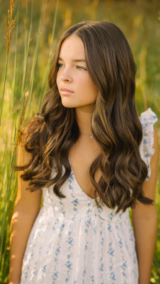 A young woman with long wavy brown hair stands in a sunlit field, wearing a white dress with blue floral patterns. She looks thoughtfully to the side, surrounded by tall grass. by Justin Salem Meyer