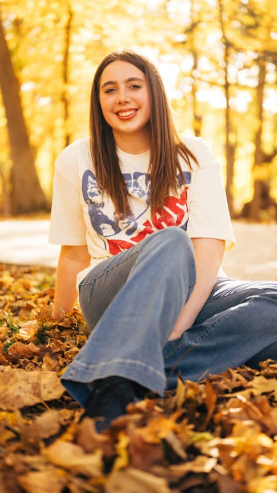 A young woman with long brown hair, wearing a graphic white T-shirt and blue jeans, sits on autumn leaves in a sunlit park, smiling at the camera. Trees with golden foliage are visible in the background. by Justin Salem Meyer