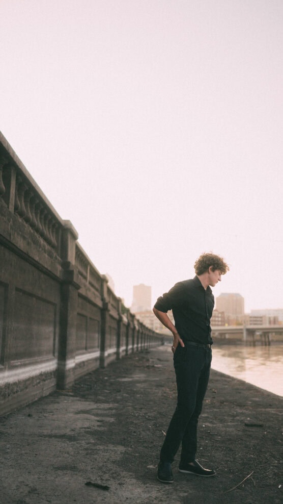 A person with curly hair, dressed in black, stands on a riverside walkway next to a concrete wall, looking down with buildings and water visible in the background at sunset. by Justin Salem Meyer