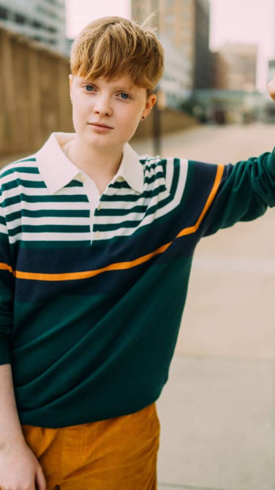 A person with short, light brown hair stands outdoors on a city sidewalk, wearing a green, white, and orange striped shirt and mustard-colored pants, looking calmly at the camera. by Justin Salem Meyer