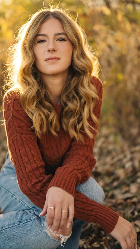 A young woman with wavy blonde hair wearing a rust-colored sweater and ripped jeans sits outdoors on fallen leaves, with golden sunlight illuminating her hair and a blurred background of autumn foliage. by Justin Salem Meyer