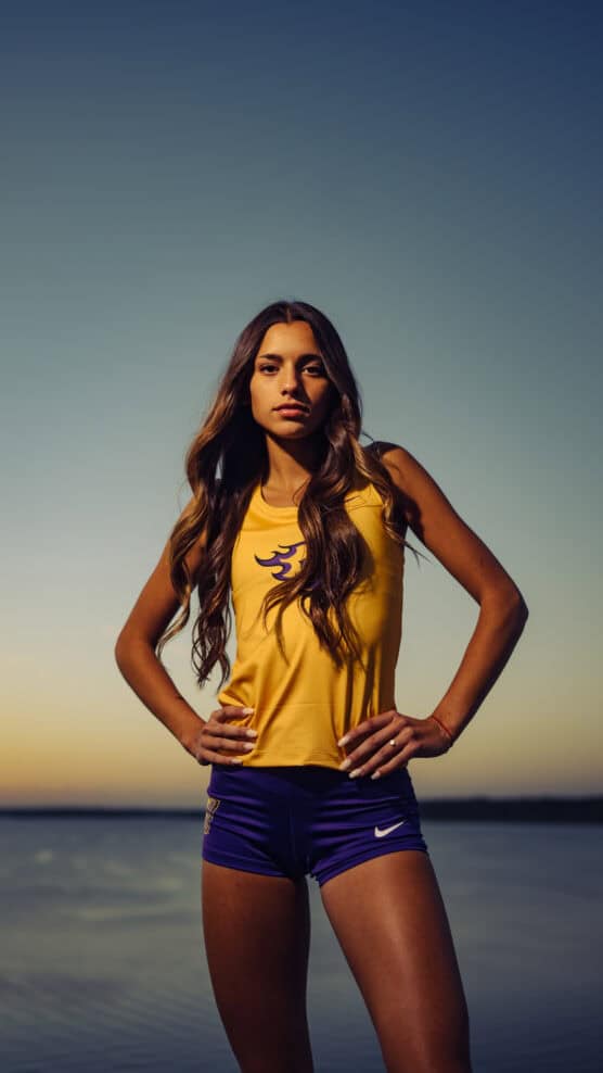 A young woman wearing a yellow athletic tank top and purple shorts stands confidently with hands on hips by a calm body of water at sunset, with a clear sky in the background. by Justin Salem Meyer