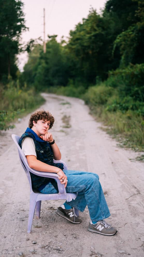 A young person with curly hair sits sideways on a plastic chair placed on a dirt road, surrounded by greenery. They wear jeans, sneakers, and a patterned vest, looking thoughtfully at the camera. by Justin Salem Meyer