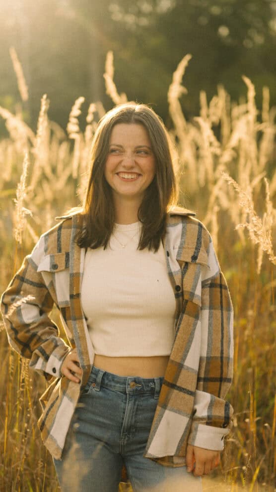 A young woman with long brown hair, wearing a plaid shirt over a white crop top and jeans, stands smiling in a sunlit field with tall grass in the background. by Justin Salem Meyer