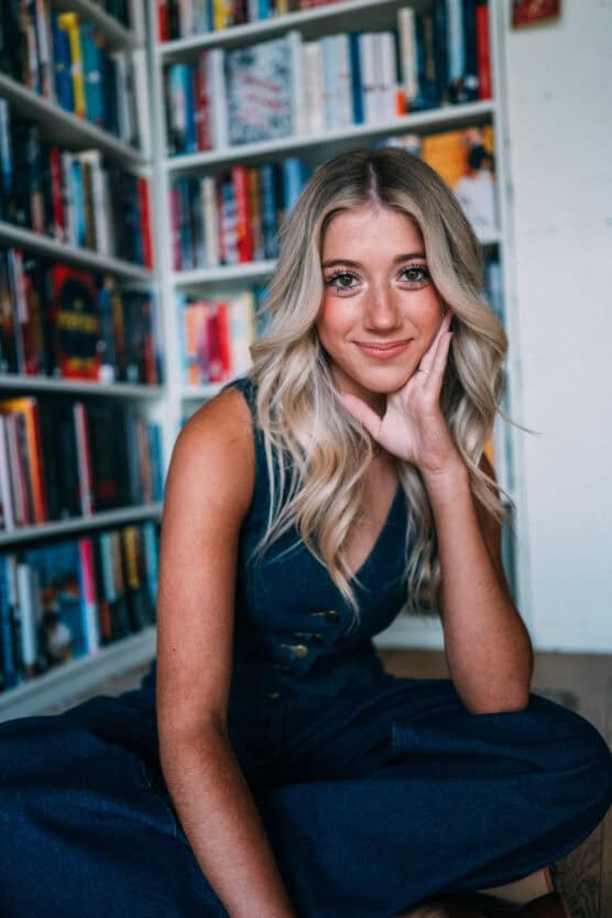 A young woman with long blonde hair and a denim outfit sits cross-legged on the floor, resting her chin on her hand, smiling softly. She is in front of colorful bookshelves filled with various books. by Justin Salem Meyer