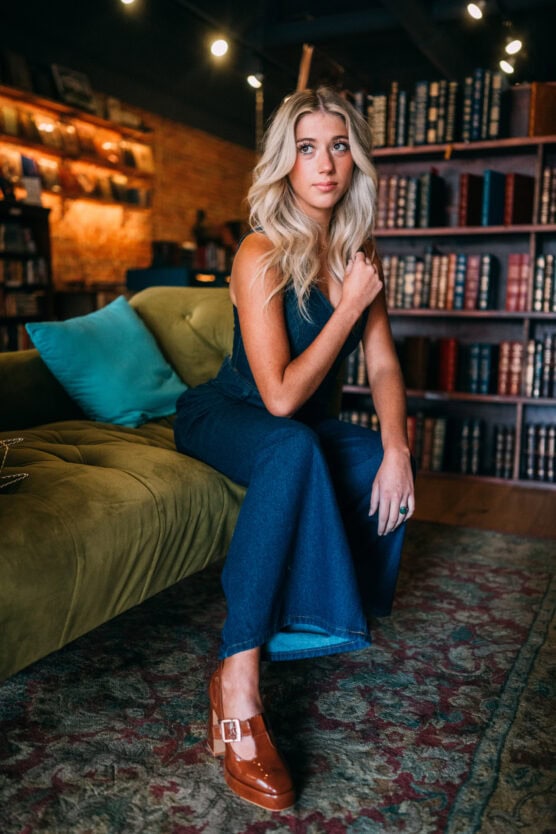 A woman with long blonde hair, dressed in a denim jumpsuit and brown shoes, sits on a green couch in a cozy library with bookshelves and warm lighting. by Justin Salem Meyer