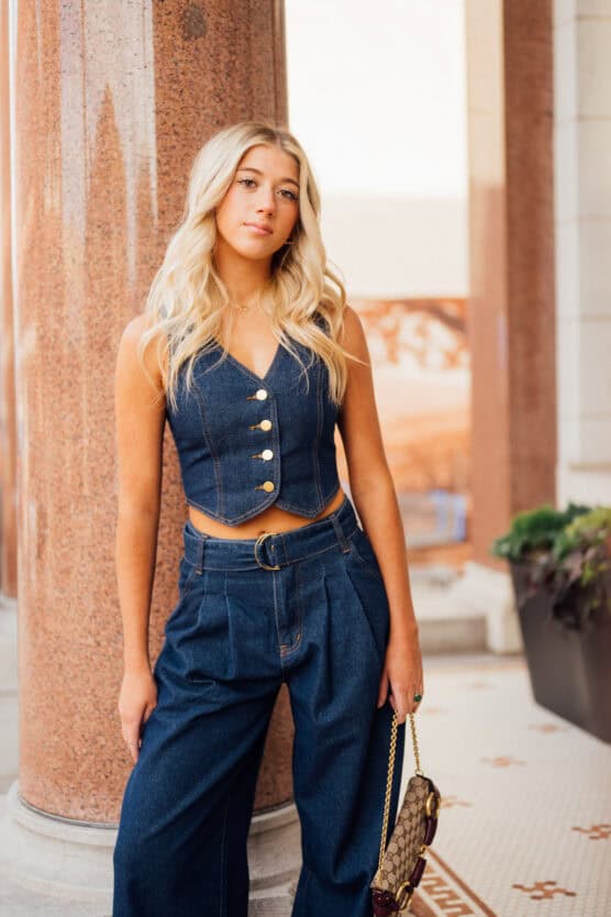 A young woman with long blonde hair stands by a marble column, wearing a sleeveless denim top with buttons and high-waisted wide-leg denim pants, holding a brown handbag and looking at the camera with a relaxed expression. by Justin Salem Meyer