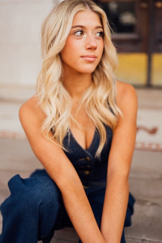 A young woman with long, wavy blonde hair sits on outdoor steps, looking to the side. She wears a sleeveless dark blue outfit and has a thoughtful expression. The background is slightly blurred. by Justin Salem Meyer