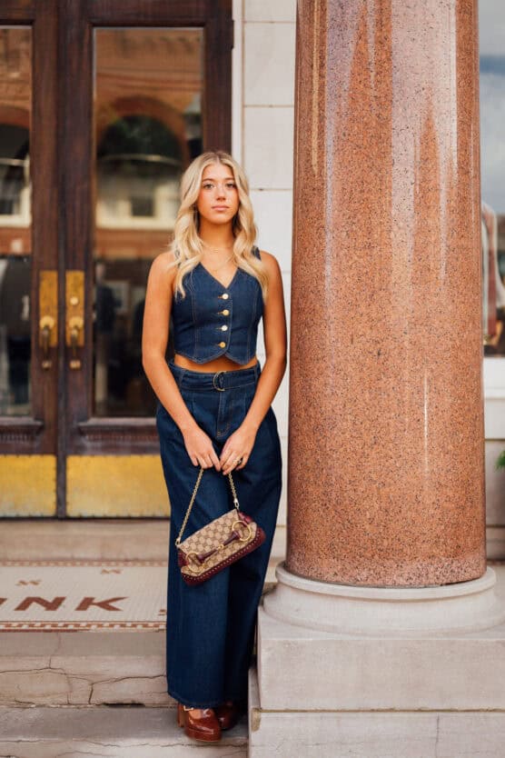 A young woman with long blonde hair stands by a large marble column, wearing a blue sleeveless denim top and wide-leg pants, holding a small purse, and looking calmly at the camera. by Justin Salem Meyer