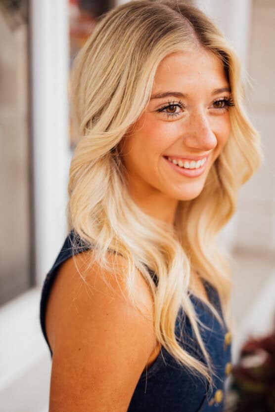 A young woman with long, wavy blonde hair and freckles smiles while looking slightly to the side. She wears a sleeveless dark top and stands in front of a blurred light background. by Justin Salem Meyer