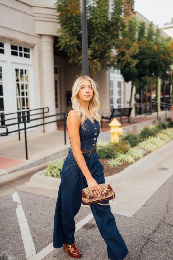 A young woman with long blonde hair walks confidently down a city street, wearing a sleeveless denim jumpsuit and brown shoes, holding a patterned handbag. Trees and storefronts line the sidewalk behind her. by Justin Salem Meyer