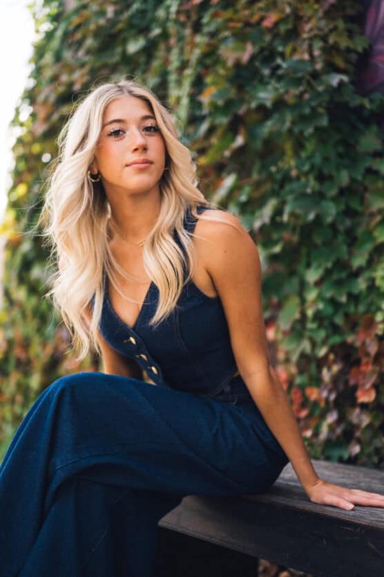A young woman with long, wavy blonde hair sits on a bench outdoors, wearing a sleeveless dark blue jumpsuit. She looks confidently at the camera, with a background of green ivy covering a wall. by Justin Salem Meyer