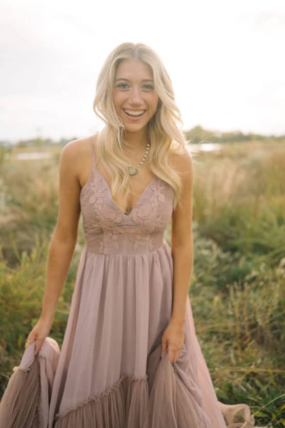 A smiling woman with long blonde hair, wearing a sleeveless lavender dress with lace details, stands outdoors in a grassy field under soft sunlight. by Justin Salem Meyer