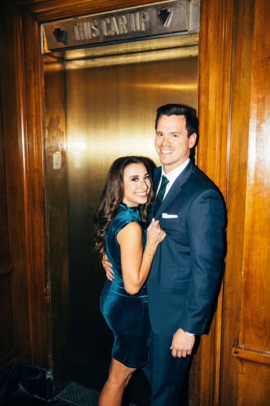 A woman in a teal velvet dress and a man in a navy suit smile while standing closely together in front of an old-fashioned brass elevator door with a sign reading This Car Up. by Justin Salem Meyer