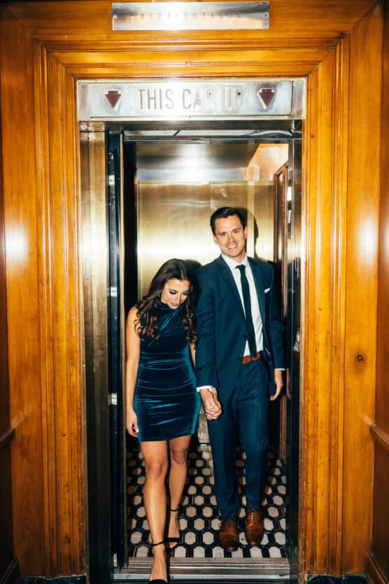 A woman in a blue velvet dress and a man in a blue suit hold hands and smile as they walk out of a vintage elevator with wood-paneled walls and a sign above that reads “THIS CAR UP.”. by Justin Salem Meyer