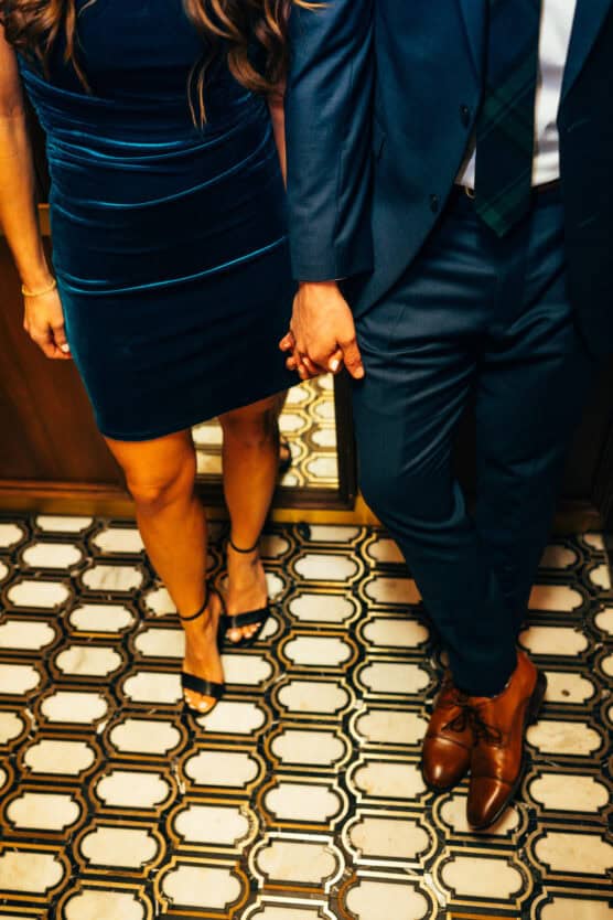 A woman in a dark velvet dress and heels holds hands with a man in a blue suit and brown shoes as they stand side by side on a patterned tile floor. Their heads are out of frame. by Justin Salem Meyer