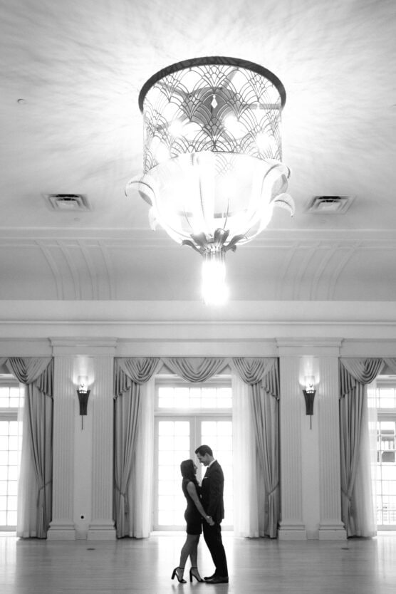 A couple stands close together in the center of an elegant, spacious room with tall windows and draped curtains. A large, ornate chandelier hangs above them, casting light across the scene. The atmosphere is romantic and serene. by Justin Salem Meyer