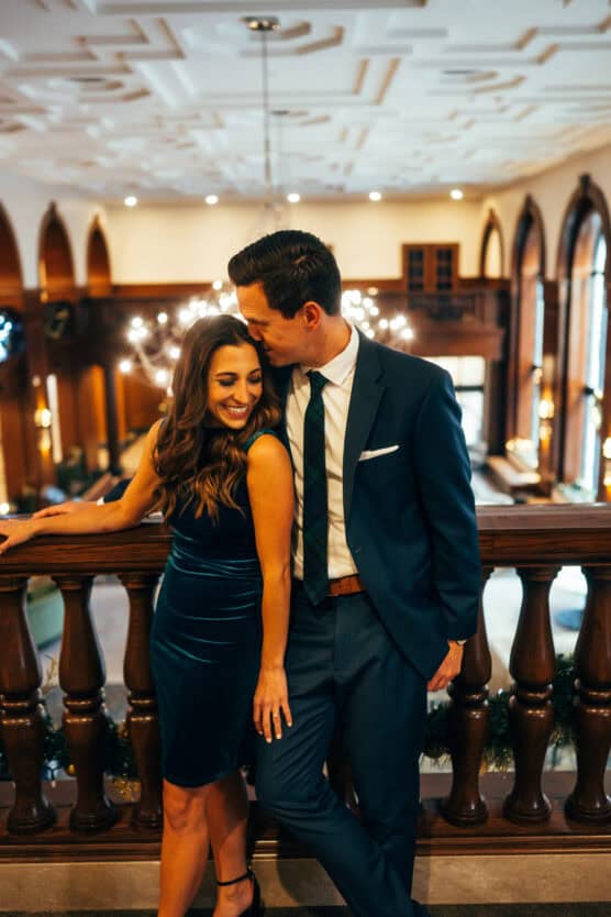 A couple dressed in formal attire stands close together, smiling and leaning on a wooden balcony railing in an elegant, warmly lit room with high ceilings and arched windows. by Justin Salem Meyer