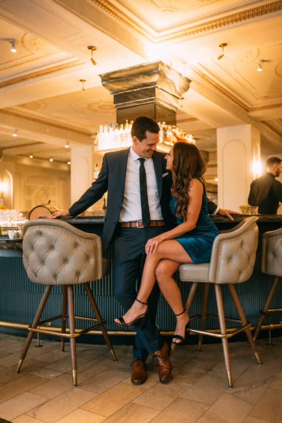 A man in a dark suit and a woman in a blue velvet dress sit closely together on barstools at an elegant bar, smiling at each other in a warmly lit, stylish setting. by Justin Salem Meyer