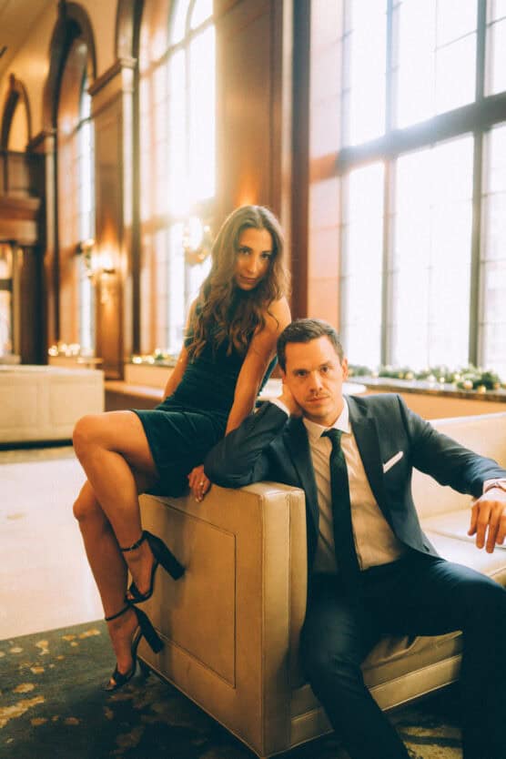 A woman in a dark dress sits on the armrest of a cream-colored chair, while a man in a suit sits beside her. They pose confidently in an elegant, sunlit room with large windows in the background. by Justin Salem Meyer
