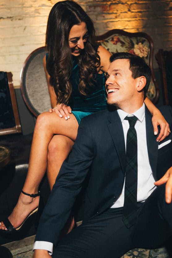 A woman in a teal dress sits on a chair, smiling and looking down at a man in a suit who is sitting on the floor, looking up at her. They appear happy and relaxed in a warmly lit room. by Justin Salem Meyer