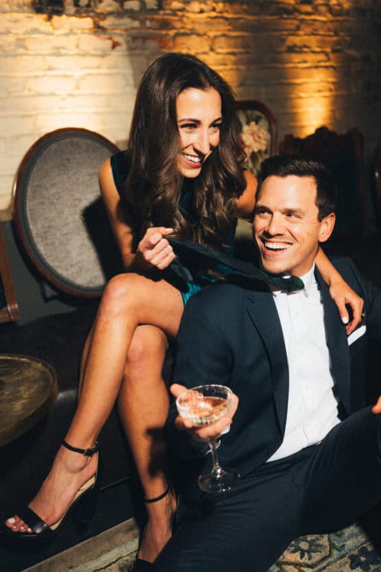 A smiling woman in a green dress sits on a couch, playfully tugging a man’s black bow tie. The man, dressed in a suit, laughs and holds a cocktail glass, both enjoying a lively moment in a warmly lit room. by Justin Salem Meyer