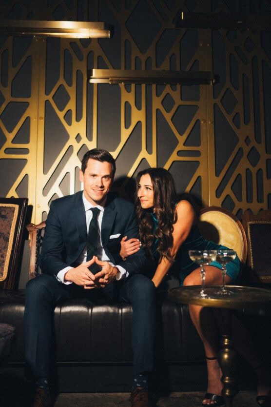 A man in a dark suit sits on a black couch, smiling, with a woman in a teal dress beside him, gently holding his arm. They are in a stylish, dimly lit room with gold geometric wall patterns. by Justin Salem Meyer