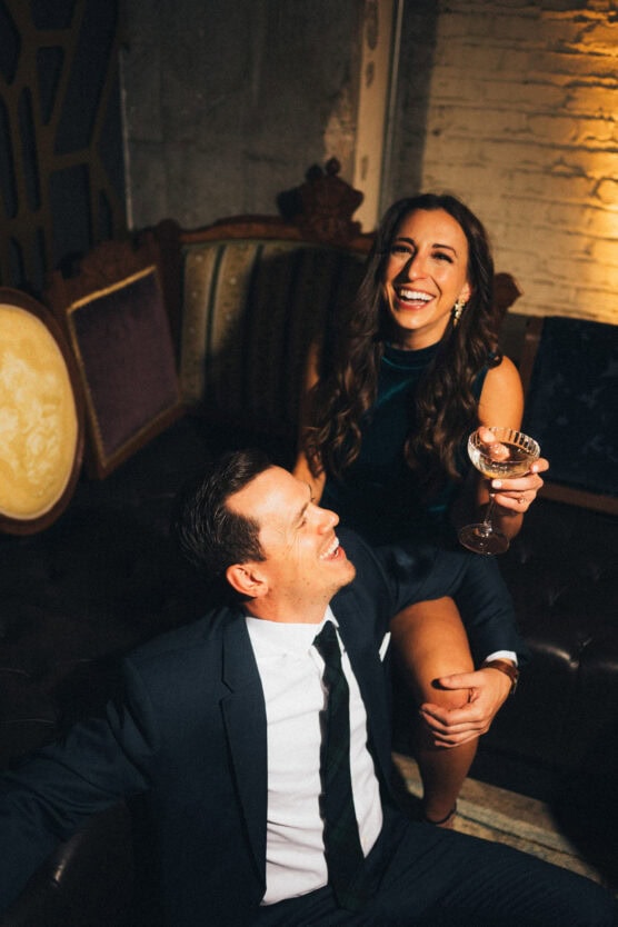 A smiling woman holding a cocktail glass sits on the arm of a couch, laughing with a man in a suit who is seated in front of her in a warmly lit, vintage-style room. by Justin Salem Meyer