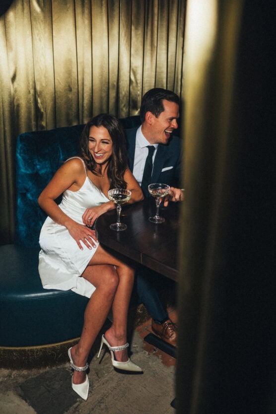 A woman in a white dress and a man in a dark suit sit closely together, smiling and holding champagne glasses at a small table in a dim, elegant setting with gold curtains. by Justin Salem Meyer