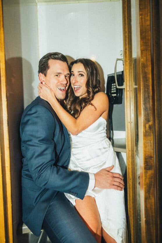 A smiling couple dressed in formal attire, with the woman in a white dress and the man in a suit, pose playfully together inside a wooden phone booth. by Justin Salem Meyer