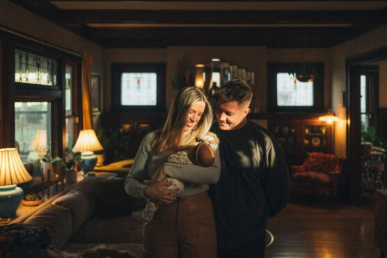 A woman and man stand together in a warmly lit living room, smiling down at a baby cradled in the womans arms. Sunlight filters in through the windows, creating a cozy, intimate atmosphere. by Justin Salem Meyer