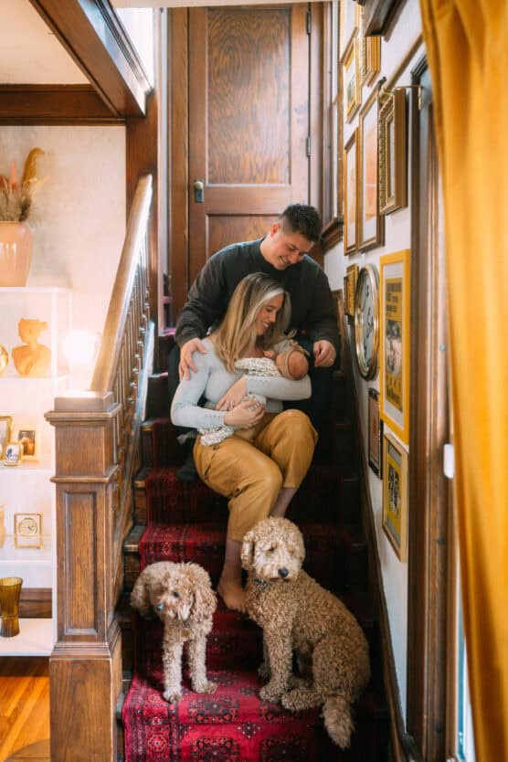 A couple sits on a carpeted staircase, holding a baby, with two curly-haired dogs sitting nearby. The walls are decorated with framed pictures and the scene is warmly lit by natural and indoor light. by Justin Salem Meyer