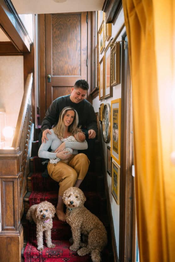 A smiling couple sits on a carpeted staircase with their baby, surrounded by framed pictures on the wall. Two small curly-haired dogs sit on the steps in front of them. Warm, cozy atmosphere. by Justin Salem Meyer