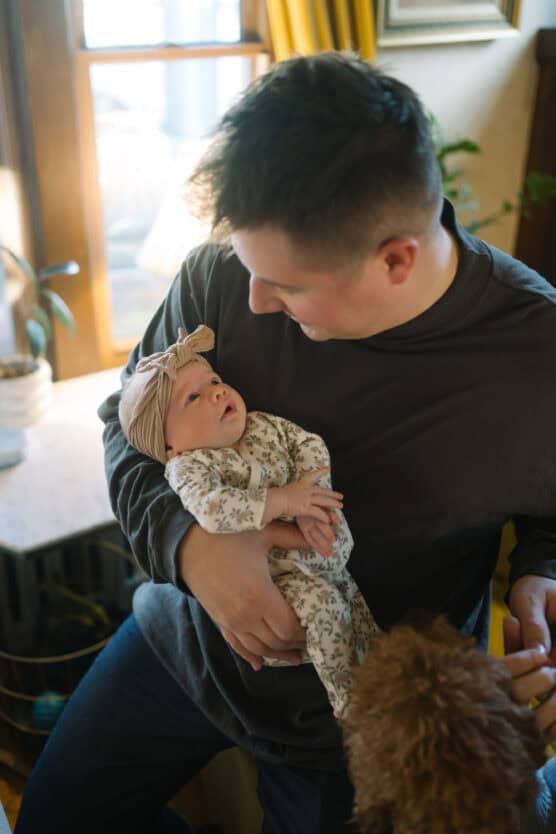 A man in a dark shirt holds a baby dressed in floral pajamas and a headband, looking up at him. They are indoors near a window, with natural light and a small dog visible at the bottom of the image. by Justin Salem Meyer