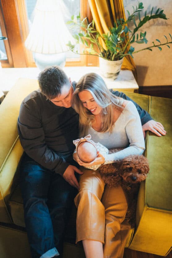 A couple sits closely on a green sofa, smiling and looking lovingly at a baby in the womans arms. A small brown dog sits beside them. A lamp and plant are in the background by a window. by Justin Salem Meyer