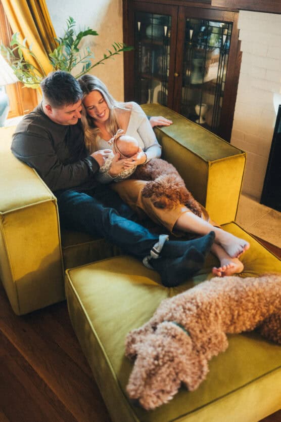 A couple sits on a yellow couch, smiling and holding a baby. A brown curly-haired dog lies beside them, while another similar dog rests on an ottoman in the foreground. The scene is cozy and warm in a home setting. by Justin Salem Meyer