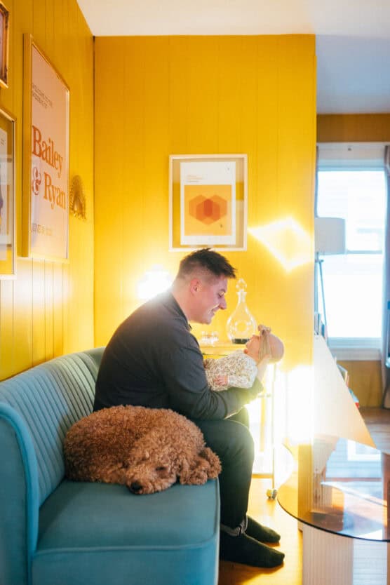 A man sits on a blue couch in a yellow-walled room, bottle-feeding a baby. A curly-haired brown dog is lying beside him. Soft sunlight streams in through a window, creating a cozy atmosphere. by Justin Salem Meyer
