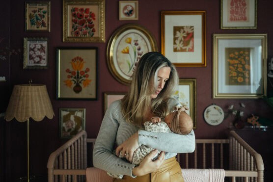 A woman with long blonde hair cradles a baby in a nursery with maroon walls, a wooden crib, and a gallery of framed floral artwork and embroidery hanging behind her. by Justin Salem Meyer