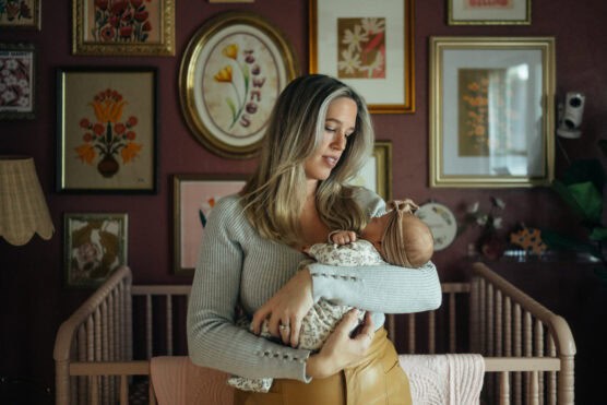 A woman with long hair holds a baby wrapped in a floral blanket and wearing a headband, standing in front of a crib and a wall decorated with framed artwork of flowers. by Justin Salem Meyer
