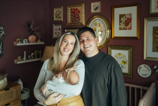 A smiling couple stands in a warmly decorated room, holding a baby wrapped in a floral blanket. The walls behind them are adorned with framed artwork and pictures. by Justin Salem Meyer