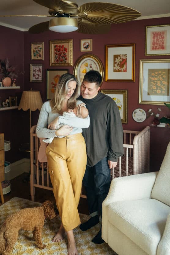 A couple stands in a cozy, art-filled nursery smiling at their newborn baby. The woman holds the baby, while the man stands close, looking at them. A small brown dog stands near their feet. by Justin Salem Meyer