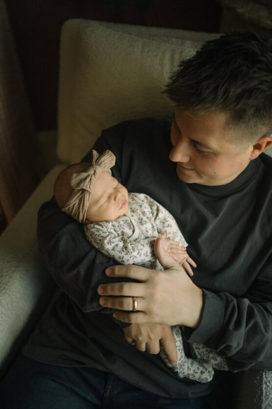 A man in a dark shirt sits in a chair, gently cradling a sleeping baby dressed in a floral onesie and headband. The man looks down at the baby with a tender expression. by Justin Salem Meyer