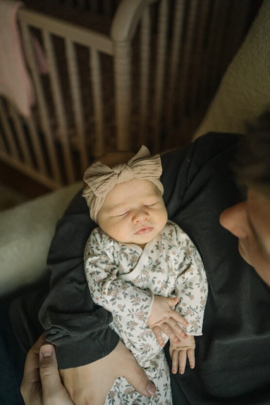 A newborn baby with a beige bow headband and floral onesie sleeps in an adults arms. The adult is wearing a dark shirt, and a crib is visible in the background. by Justin Salem Meyer