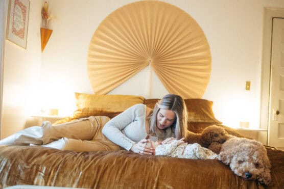 A woman lies on a bed with brown bedding, gently holding a baby. Two curly-haired dogs are also lying on the bed beside her. The room is softly lit, and a large decorative fan hangs above the bed. by Justin Salem Meyer